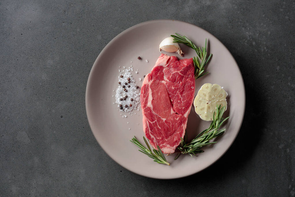 Raw beef steak for grilling with rosemary, garlic, salt, and pepper. Grey concrete background. Top view. - Foto, Imagen