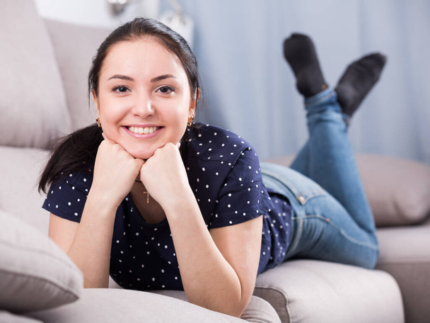 Giovane ragazza dagli occhi marroni con sorriso appoggiato sui pugni sul divano - Foto, immagini