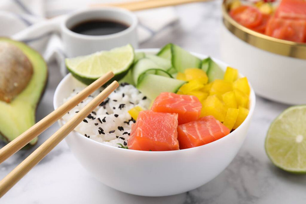 Delicious poke bowl with salmon, lime and vegetables on white marble table, closeup - Zdjęcie, obraz