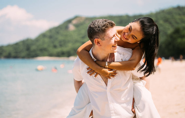 Νεαρό ευτυχισμένο ζευγάρι ανδρών και γυναικών σε λευκά ρούχα στην παραλία πορτρέτο, Girlfriend να πάρει piggyback βόλτα από το φίλο κατά την ανατολή του ηλίου πάνω από θάλασσα ωκεανό εξωτερική το καλοκαίρι ημέρα - Φωτογραφία, εικόνα
