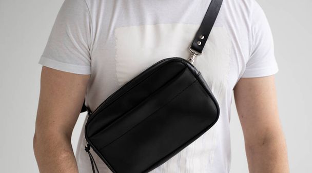 Man in a white t-shirt with a leather black handmade bag over his shoulder. Dark designer banana bag. Comfortable small bag for walking - Foto, imagen