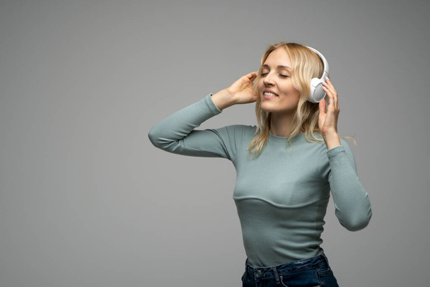 Beautiful attractive young blond woman wearing blue t-shirt and glasses in white headphones listening music and smiling on grey background in studio. Relaxing and enjoying. Lifestyle - Fotoğraf, Görsel