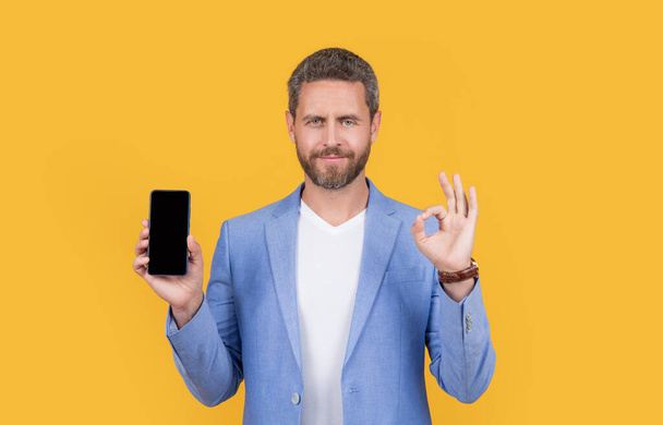 businessman hold portable phone, ok. businessman showing portable phone. businessman presenting screen of portable phone with copy space. businessman with portable phone isolated on yellow. - Foto, Imagem