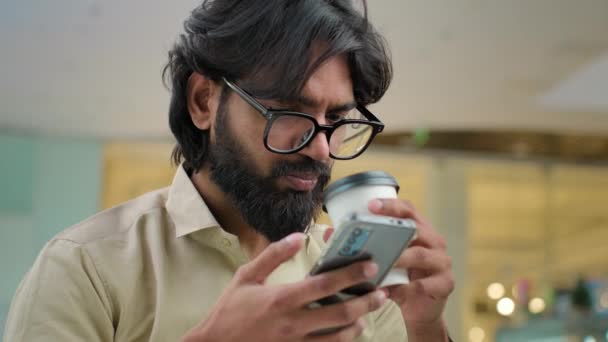 Close up smiling Indian bearded millennial man in glasses drinking coffee scrolling browsing using smartphone in cafe. Arabian businessman drink tea use mobile phone app for business chatting online - Filmagem, Vídeo