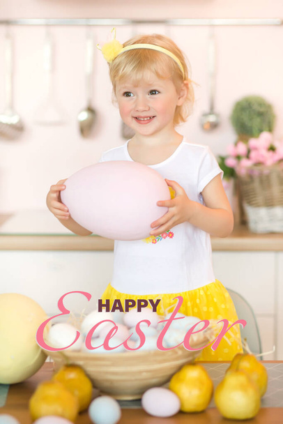 Osterkarte. Ein kleines süßes Mädchen lächelt mit einem großen Osterei in der heimischen Küche neben einem Korb pastellfarbener Ostereier - Foto, Bild