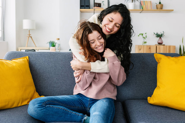 Две многорасовые молодые женщины смеются и веселятся вместе дома. Улыбающаяся латиноамериканка обнимается со своей подружкой, сидя на диване. Концепция дружбы - Фото, изображение