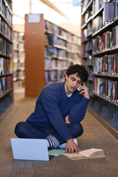 Ισπανόφωνος multitasking έξυπνος φοιτητής μιλώντας στο κινητό τηλέφωνο, διαβάζοντας και ψάχνοντας πληροφορίες για ένα βιβλίο, ενώ κάθεται στο φορητό υπολογιστή στο πάτωμα, στην κατασκήνωση βιβλιοθήκη.Εκπαίδευση και οι άνθρωποι έννοια - Φωτογραφία, εικόνα