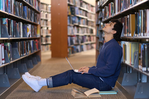Σκεπτικό multitasking Ισπανόφωνος νεαρός άνδρας, φοιτητής ακουμπά στη βιβλιοθήκη με τη λογοτεχνία, χρησιμοποιώντας φορητό υπολογιστή, γράφει κείμενο, κάνει έρευνες, ενώ μελέτες για τον ελεύθερο χρόνο του, κάθεται στο πάτωμα σε μια πανεπιστημιούπολη βιβλιοθήκη - Φωτογραφία, εικόνα
