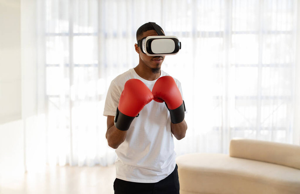 Jonge zwarte man vechter in VR-headset en rode bokshandschoenen met virtuele bokstraining thuis. Afro-Amerikaanse man training via augmented reality ervaring, kopieer ruimte - Foto, afbeelding