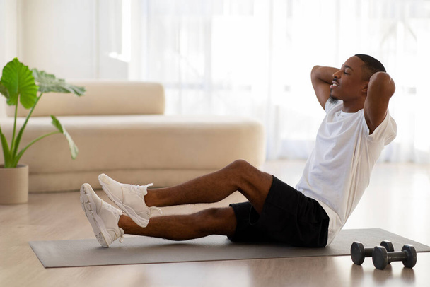 Motivated athletic handsome young african american man in sportswear sitting on yoga mat, doing crunches for abs workout at home, using fitness tools, looking at copy space, side view - Foto, Bild