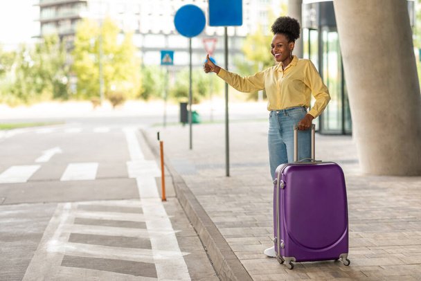 African American Tourist Lady Stopping Taxi With Thumbs Up Gesture Standing With Travel Suitcase Near Modern Airport Outdoor. Female Traveler Hailing A Cab. Transportation Concept - 写真・画像