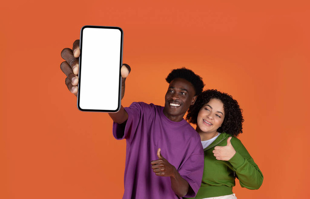 Leuke vrolijke multiraciale jonge paar hipsters tonen gloednieuwe mobiele telefoon met wit leeg scherm en duim omhoog op de camera, aanbevelen mooie dating app, geïsoleerd op rode achtergrond, mockup - Foto, afbeelding