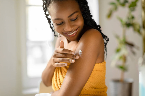 Έννοια φροντίδας σώματος. Ευτυχισμένο μαύρο γυναίκα εφαρμογή ενυδατική λοσιόν στον ώμο, χρησιμοποιώντας θρεπτική κρέμα μετά το μπάνιο, απολαμβάνοντας θεραπείες του δέρματος στο σπίτι - Φωτογραφία, εικόνα