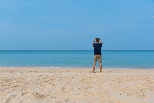 Un hombre adulto asiático mayor usando un teléfono inteligente tomando fotografías en la playa. - Foto, imagen