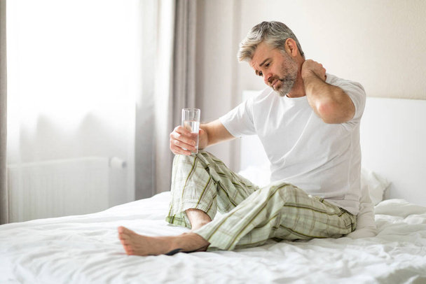 Tired middle aged man in pajamas suffering from neck pain after sleeping in bedroom, waking up in morning, sitting on bed, touching his neck, taking painkiller, experiencing body aches, copy space - Fotoğraf, Görsel