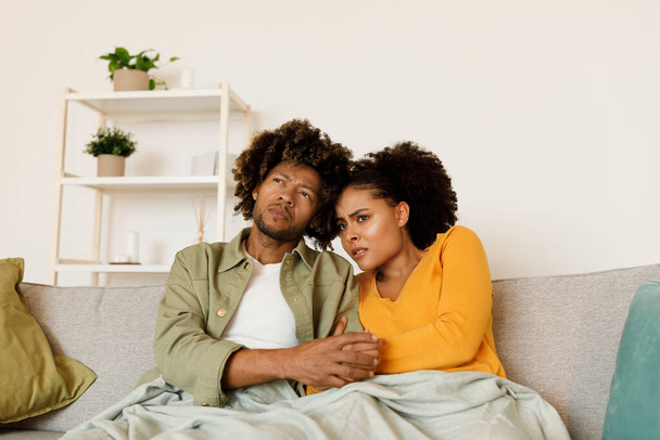 Беспокойная афро-американская пара смотрит жуткий фильм или негативные новости по телевизору, обнимаясь на диване вместе в современной гостиной дома. Критика и разочарование - Фото, изображение