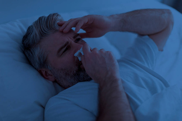 Cold, flu, coronavirus. Sick unhappy handsome mature man awake at night, suffering from runny nose, lying in bed in darkness, cleaning nose with napkin and touching his head, closeup shot - Photo, Image