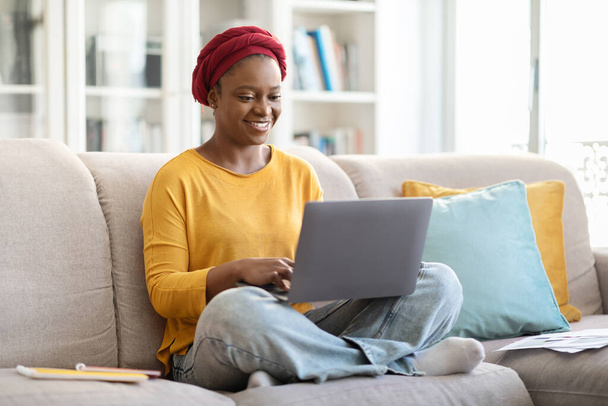 Cheerful pretty millennial african american woman with red turban in casual outfit sitting on couch, typing on pc laptop keyboard and smiling, female digital nomad working from home, copy space - Foto, afbeelding