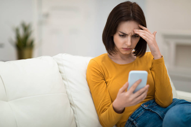 Confused pensive upset brunette young woman in casual sitting on couch at home, looking at smartphone screen and touching her face, reading weird email or message, copy space - Foto, imagen
