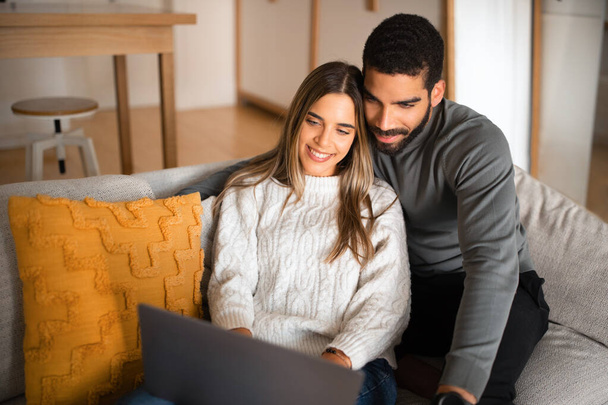 Lächelnder arabischer Millennial-Typ mit Bart umarmt europäische Dame, schaut Video auf Laptop, sitzt auf Couch im Wohnzimmer Interieur. Unterhaltung und Online-Telefonate zu Hause in der Freizeit, soziale Netzwerke und Blog - Foto, Bild