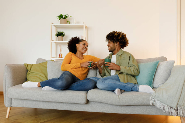 Ευτυχισμένο παντρεμένο μαύρο ζευγάρι που μιλάει και πίνει καφέ καθισμένος στον καναπέ στο σπίτι τα σαββατοκύριακα. Σύζυγοι χαμογελώντας ο ένας στον άλλο απολαμβάνοντας συνομιλία κρατώντας κούπες στο άνετο σαλόνι - Φωτογραφία, εικόνα