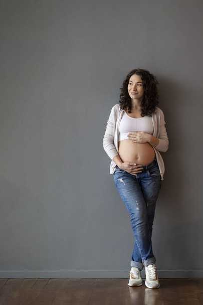 Full Length Shot Of Beautiful Young Pregnant Woman Embracing Belly And Looking Aside, Happy Smiling Female Expecting Baby Posing Against Grey Wall Indoors, Enjoying Healthy Pregnancy, Copy Space - Foto, immagini