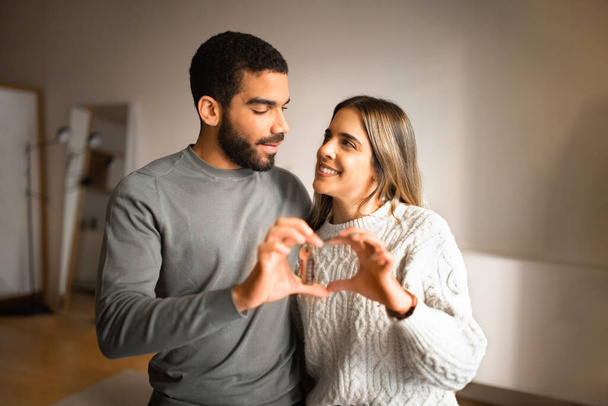 Smiling millennial european female and arab guy have fun, make heart sign with hands together, hold keys in room interior. Love of relationships at home, real estate, buy new apartment and rent house - Foto, Imagem