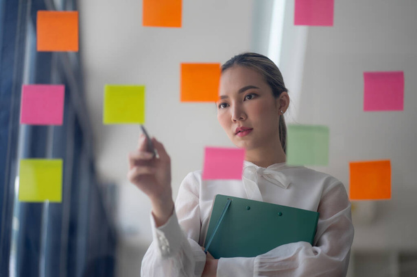 Una donna d'affari usa note post-it per condividere idee, mostrando il concetto di brainstorming. Le note adesive sono posizionate su una parete di vetro - Foto, immagini