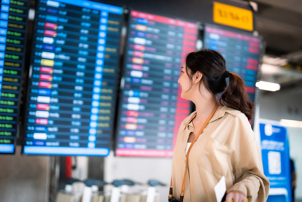 Fiatal ázsiai nő útlevéllel és beszállókártyával segítségül a nemzetközi repülőtéren nézi a repülési információs táblát, ellenőrzi a járatát - Fotó, kép