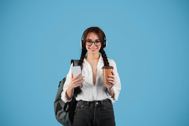 Cheerful european teen girl with pigtails, glasses, backpack in wireless headphones hold phone and cup of takeaway coffee isolated on blue background. Take break with audio app, study and education - Photo, image