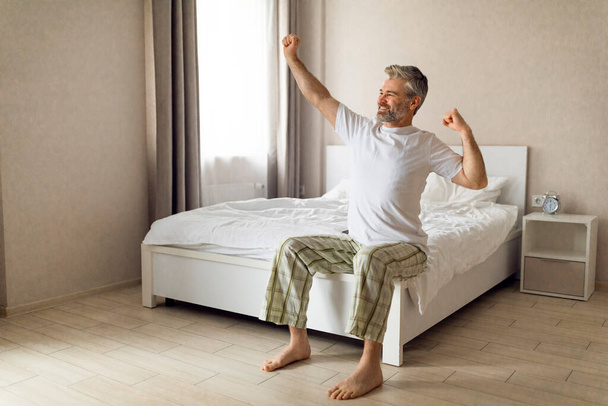 Fröhlich fröhlicher gutaussehender grauhaariger europäischer Mann mittleren Alters sitzt auf dem Bett und streckt die Arme aus, schaut zum Fenster und lächelt, genießt den neuen sonnigen Tag, den Kopierraum. Gutes Schlafkonzept - Foto, Bild