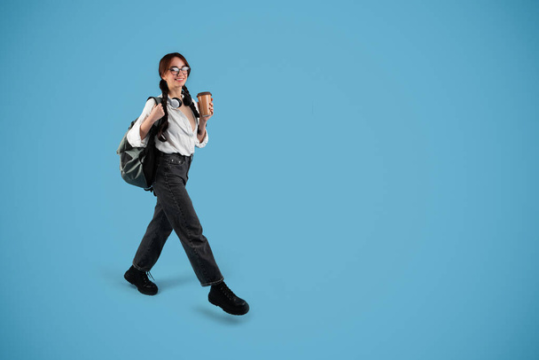 Glad european teen female student with pigtails, glasses, backpack and wireless headphones hold cup of takeaway coffee, walk, isolated on blue background, full length. Study and education, ad, offer - Fotografie, Obrázek
