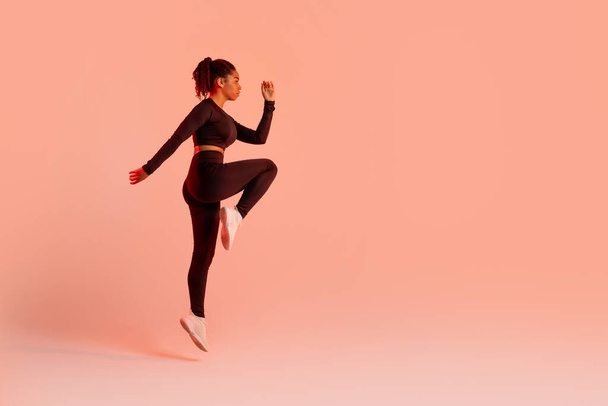 Fit athlete woman jumping while exercising over peach neon studio background, looking aside at free space, full length, side view. Fitness workout and sport motivation concept - Photo, Image