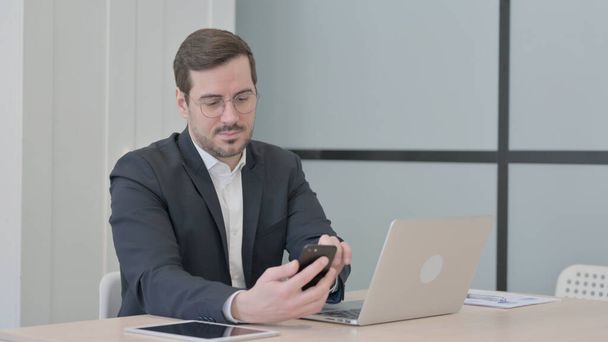 Liikemies käyttää älypuhelinta, kun käytät kannettavaa tietokonetta - Valokuva, kuva