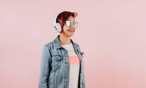 young hispanic woman girl listening music with headphones  and holding mobile phone on coral pink background with copy space portrait in Mexico Latin America - Foto, imagen