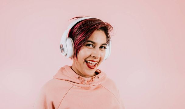 young hispanic woman girl listening music with headphones  and holding mobile phone on coral pink background with copy space studio portrait in Mexico Latin America - Foto, immagini