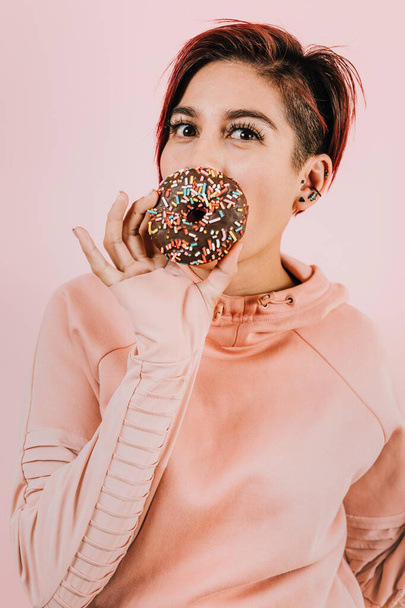 Portrait of young hispanic woman eating chocolate donuts on coral pink background in Mexico Latin America - 写真・画像