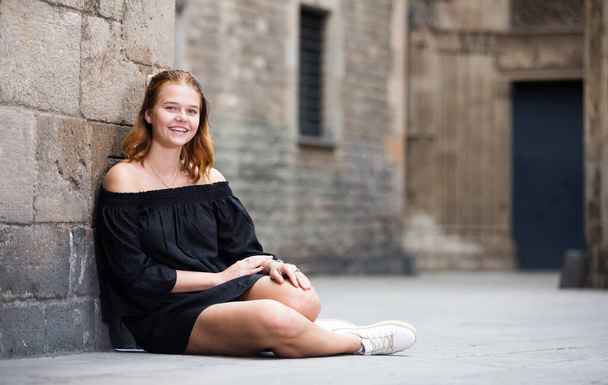 Positive young girl sitting near the stone wall at the street - Foto, Bild