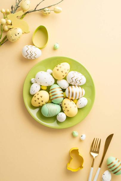 Easter atmosphere concept. Top view vertical photo of green plate with lot of colorful easter eggs cutlery baking molds and easter plant on isolated pastel beige background - Фото, изображение