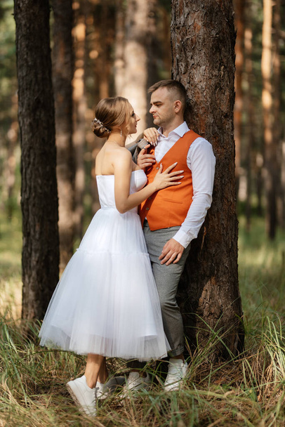 joven pareja novia en un vestido corto blanco y novio en un traje gris en un bosque de pinos entre los árboles - Foto, Imagen