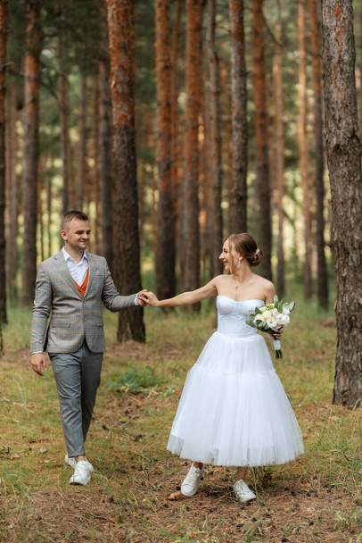 young couple bride in a white short dress and groom in a gray suit in a pine forest among the trees - Zdjęcie, obraz