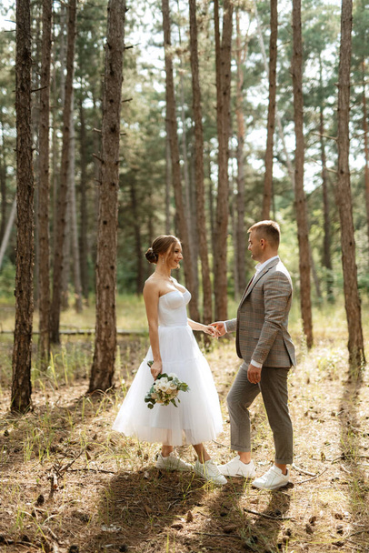 jong stel bruid in een witte korte jurk en bruidegom in een grijs pak in een dennenbos tussen de bomen - Foto, afbeelding