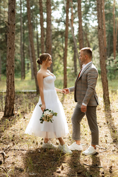 fiatal pár menyasszony egy fehér rövid ruha és a vőlegény egy szürke öltöny egy fenyőerdőben a fák között - Fotó, kép