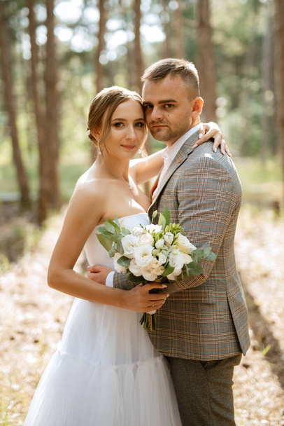 jeune couple mariée dans une robe courte blanche et marié dans un costume gris dans une forêt de pins parmi les arbres - Photo, image