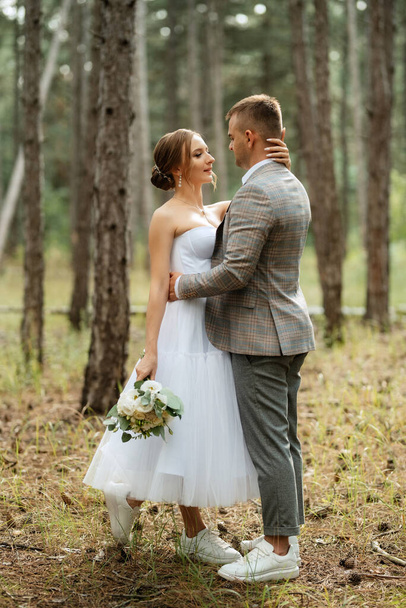 young couple bride in a white short dress and groom in a gray suit in a pine forest among the trees - Photo, image