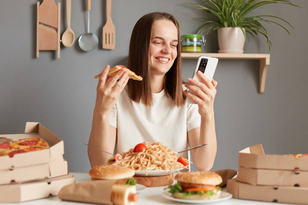 Image of smiling happy woman with brown hair wearing white t shirt sitting in kitchen at table, eating pizza and using cell phone, checking social networks while having snack. - 写真・画像