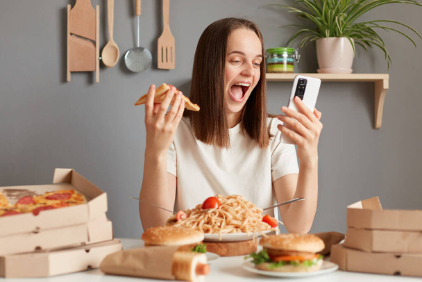Extremely happy overjoyed Caucasian woman with brown hair wearing white t shirt sitting in kitchen at table and holding slice of pizza in hands, having dinner with junk food, using mobile phone. - Foto, Bild