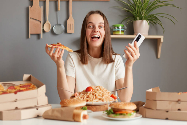Indoor shot of amazed excited young adult woman with brown hair wearing white t shirt sitting in kitchen at table and looking at camera, holding in hands, smart phone and slice of pizza. - Fotoğraf, Görsel
