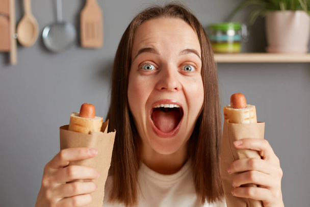 Surprised wondered woman keeps mouth widely opened holds appetizing hot dogs, eating fast food, having cheat meal and unhealthy nutrition, screaming with amazement. - Fotografie, Obrázek