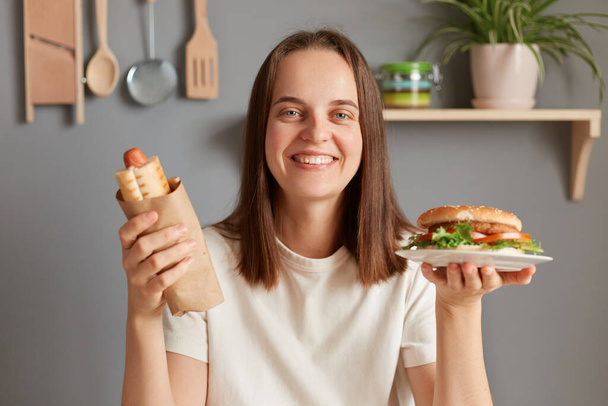 Indoor shot of woman sitting in kitchen and holding hot dog ans sandwich, looking smiling at camera, enjoying eating junk food, looking at camera with toothy smile. - Photo, Image